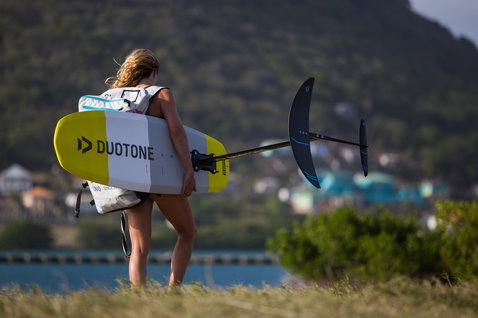 Duotone Pace 2021 Foil-Kiteboard beim Foilen an einer grasbewachsenen Küste mit einer Siedlung am Wasser in der Ferne