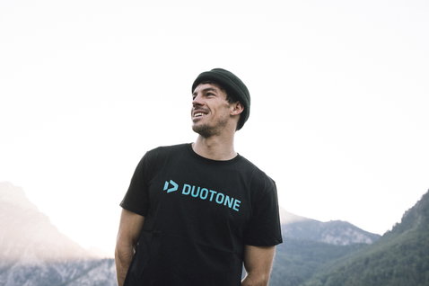 Camiseta de manga corta en negro con logo en verde azulado en pose de pie en paisaje de montaña