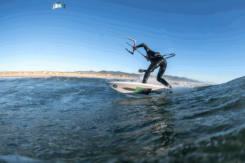Duotone Wam SLS Wave-Kiteboard beim Wellenreiten am Ozeanstrand mit Küste und Bergen im Hintergrund