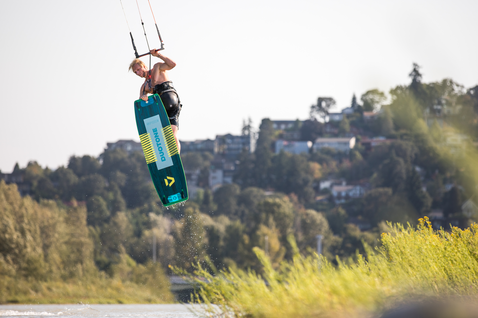 Duotone Spike twin-tip kiteboard durante il kitesurf su un lago interno con colline boscose