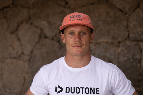 Gorra de béisbol en naranja terracota con azul marino y blanco, llevada por un modelo frente a una pared de piedra exterior