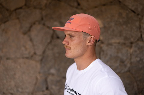 Gorra en coral salmón con azul marino y blanco, llevada por un modelo frente a una pared de piedra exterior