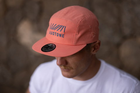 Gorra de 5 paneles Duotone en salmón coral con bordado azul marino, usada por un modelo junto a una pared de piedra exterior