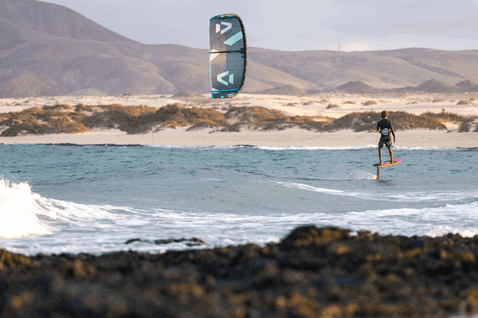 Duotone Neo SLS LEI kite durante el surf de olas en una playa oceánica con costa rocosa y dunas de arena
