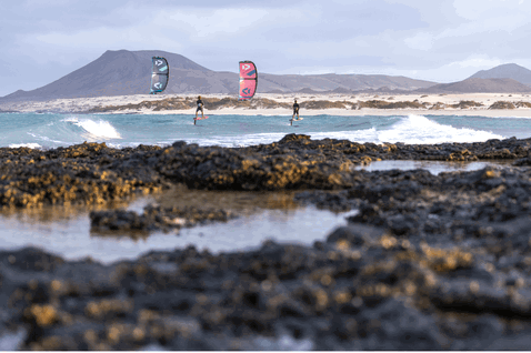 Duotone Pace planche de kite foil lors de foiling sur une côte océanique avec roches volcaniques et plage de sable avec montagnes volcaniques en fond