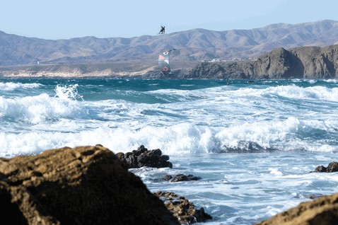 Duotone Rebel SLS LEI kite during kitesurfing at rocky coastal cliffs and ocean beach in dark charcoal, steel blue and orange