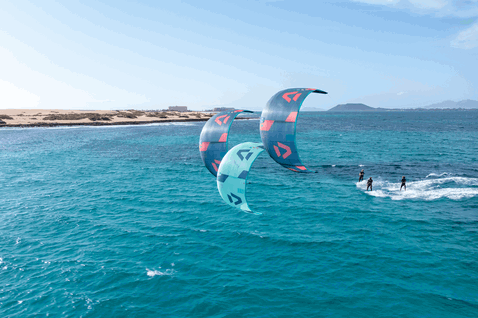 Duotone Neo LEI kite durante il surf sulle onde su una spiaggia oceanica con dune costiere in blu verde acqua e corallo