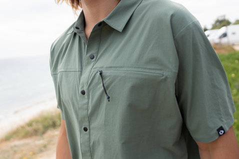 Duotone Apparel Shirt Wave Kurzarmhemd in dusty olive, getragen an Küstenklippe mit Meer und Strand im Hintergrund