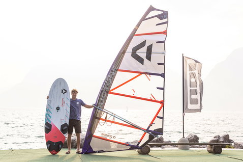 Planche de windsurf Duotone Falcon Fin D/LAB en pose debout au bord d'un lac ou de la mer avec des montagnes en arrière-plan