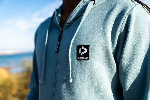 Zip-up hoodie in light teal blue with white and black, worn by a model on the coast with sea and scrubland in the background