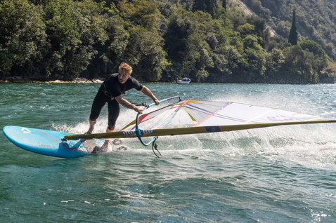 Duotone Blast SLS Windsurf-Board beim Windsurfen auf einem See mit bewaldeten Hügeln