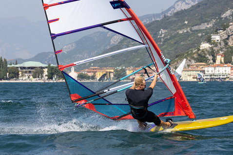 Duotone Blast SLS Windsurfboard beim Windsurfen auf einem See mit Stadt und Bergen im Hintergrund