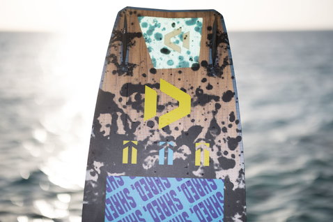 Duotone Shred kiteboard twin-tip en gris anthracite, turquoise menthe, jaune et bleu au bord de l'océan