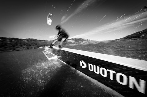 Duotone TS Park twin-tip kiteboard 151cm lors de wakestyle dans un parc câble avec montagnes en arrière-plan