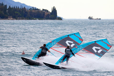 Duotone Warp_Fin race sail 7.8m during windsurfing on a lake with forested hillside and buildings in the background