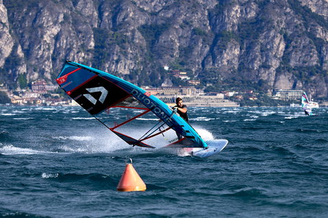 Duotone Warp_Fin race sail 7.8m² in steel blue during windsurfing on a lake with mountain backdrop