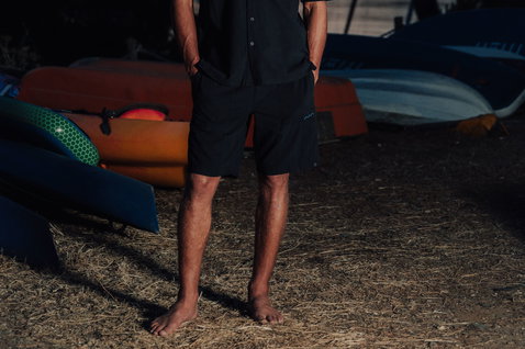 Duotone Apparel Shorts True Beach men short de plage en pose sur un bord de mer avec bateaux et herbe sèche