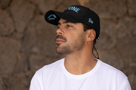 Duotone cap in black with teal accents, worn by a model at a rocky coastal location