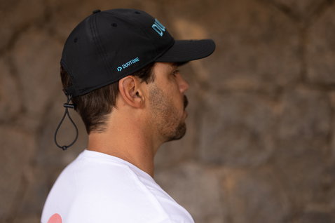 Duotone snapback cap in black with teal logo lettering, worn by a model at an outdoor stone wall