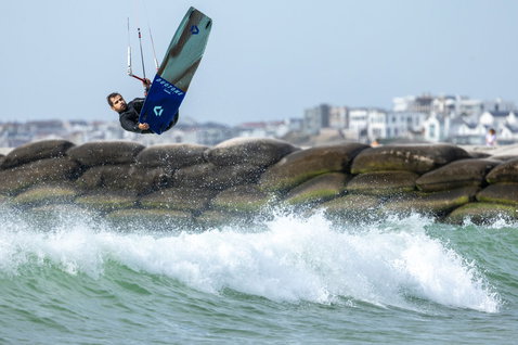 Duotone Spike Concept Blue twin-tip kiteboard during kitesurfing at a rocky coastal shore with ocean waves and town in the background