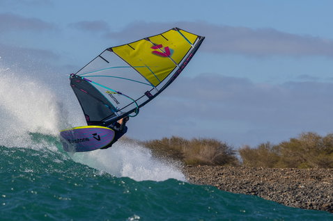 Duotone Grip [Tri] SLS windsurf board during wave riding at an ocean beach with rocky shoreline and scrubland