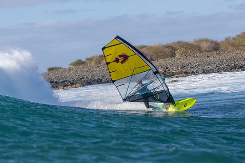 Duotone Grip [Tri] SLS windsurf board during wave riding at a rocky ocean coastline in teal