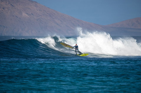 Duotone Ultra Grip [Quad] windsurf board during wave riding at an ocean wave break with arid mountain backdrop