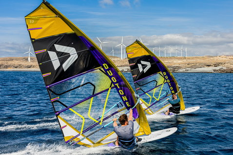 Duotone Warp_Fin voile de course 7,7 m² en windsurf sur une côte océanique avec parc éolien en arrière-plan