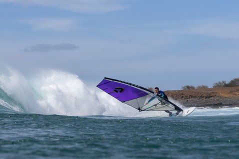 Duotone Grip [Quad] D/LAB windsurf board during wave riding at an ocean coastal break with rocky shoreline
