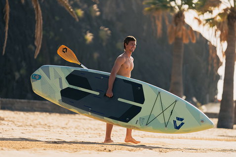 Planche de SUP gonflable Duotone Blitz Air SLS portée sur une plage ensoleillée bordée de palmiers