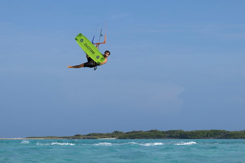 Tabla de kitesurf twin-tip Duotone Jaime Concept Blue durante un salto aéreo sobre una laguna turquesa en verde lima