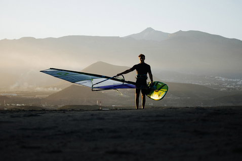 Duotone Idol freestyle windsurf sail held by rider on volcanic beach at golden hour with mountain backdrop